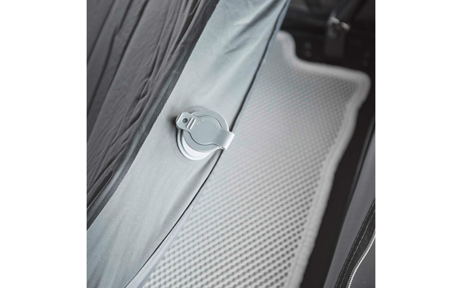 Nahaufnahme eines grauen Stoffteils mit runder weißer Kunststoffhalterung mit Clip, daneben eine hellgraue, genoppte Antirutschmatte und der Rand eines schwarzen Polstersitzes