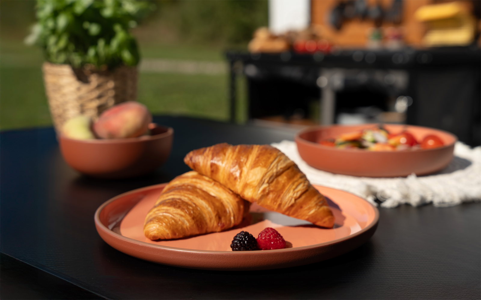 Zwei Croissants auf einem runden Teller, davor eine Brombeere und eine Himbeere; im Hintergrund Schalen mit Obst und eine Pflanze
