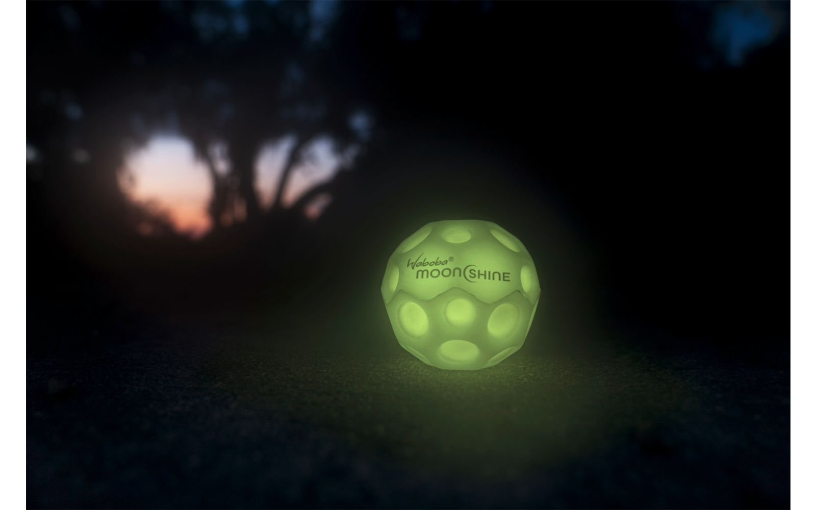 Leuchtender grüner Ball auf dunklem Boden bei Nacht mit unscharfem Baum im Hintergrund