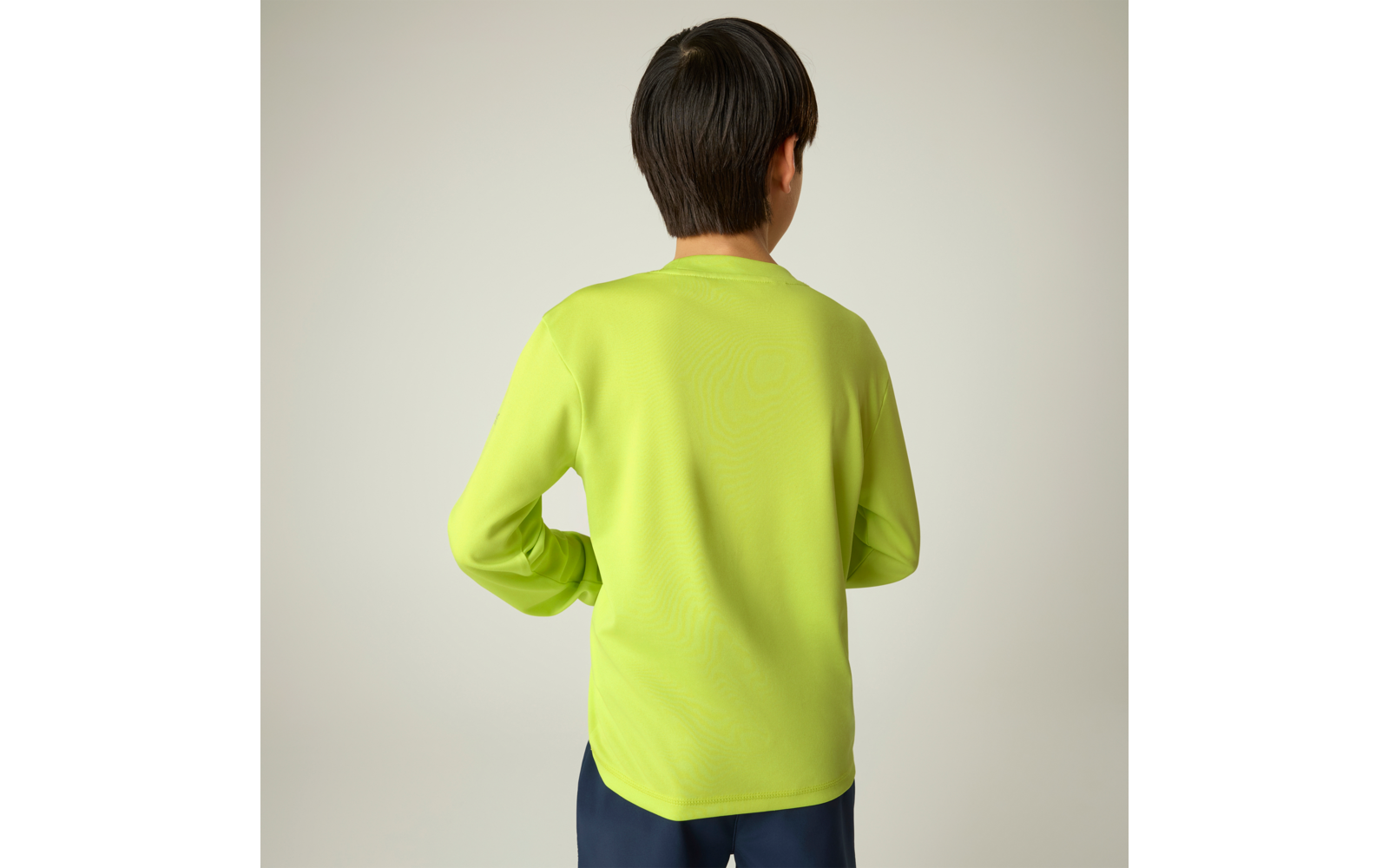 Person von hinten mit kurzem dunklem Haar, trägt ein neongrünes Langarmshirt und eine dunkle Hose vor hellem Hintergrund