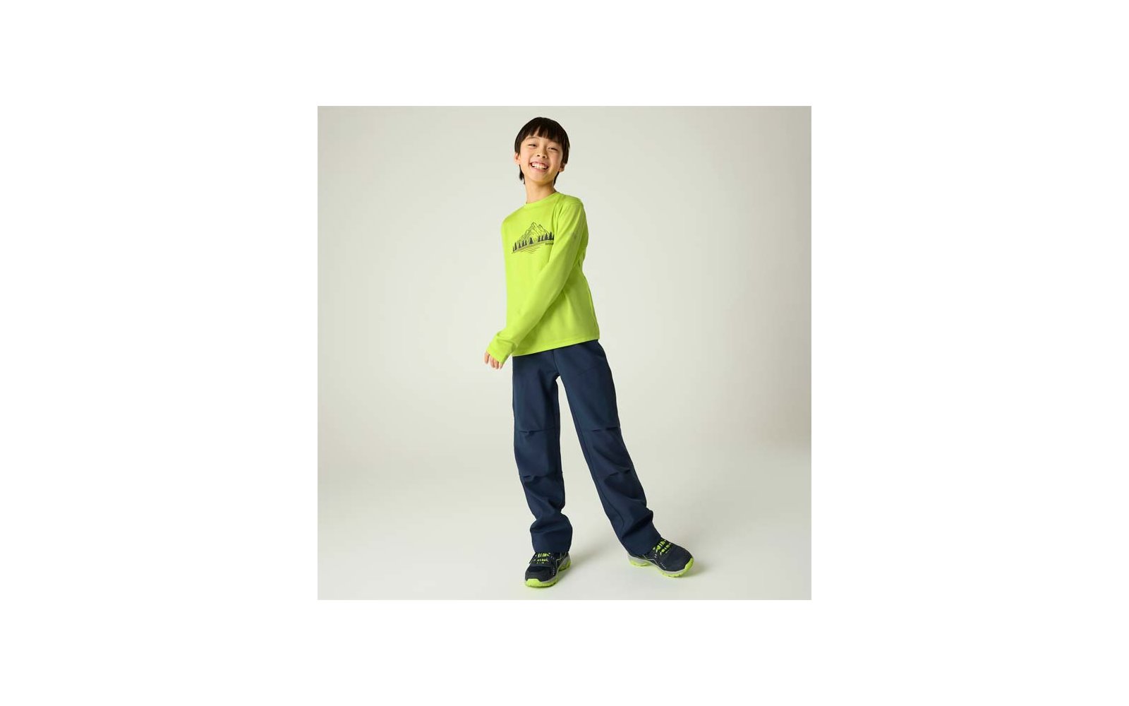 Eine lächelnde Person steht vor hellem Hintergrund, trägt ein leuchtend grünes Langarmshirt mit Berg-Motiv, dunkelblaue weite Hosen und grün-schwarze Sportschuhe