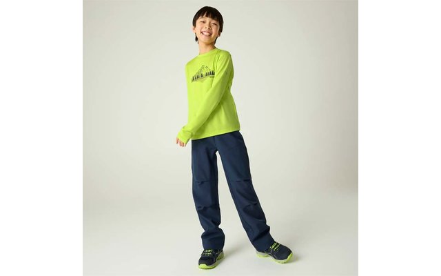 Eine lächelnde Person steht vor hellem Hintergrund, trägt ein leuchtend grünes Langarmshirt mit Berg-Motiv, dunkelblaue weite Hosen und grün-schwarze Sportschuhe