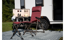 Zwei rote Klappstühle (ein hoher, ein niedriger Hocker) auf einem gemusterten Teppich vor einem weißen Wohnmobil; rechts ist ein Rad des Wohnmobils sichtbar, links ein kleiner Klapptisch mit offener Holzkiste mit Flaschen und Bechern, Gras und Bäume 