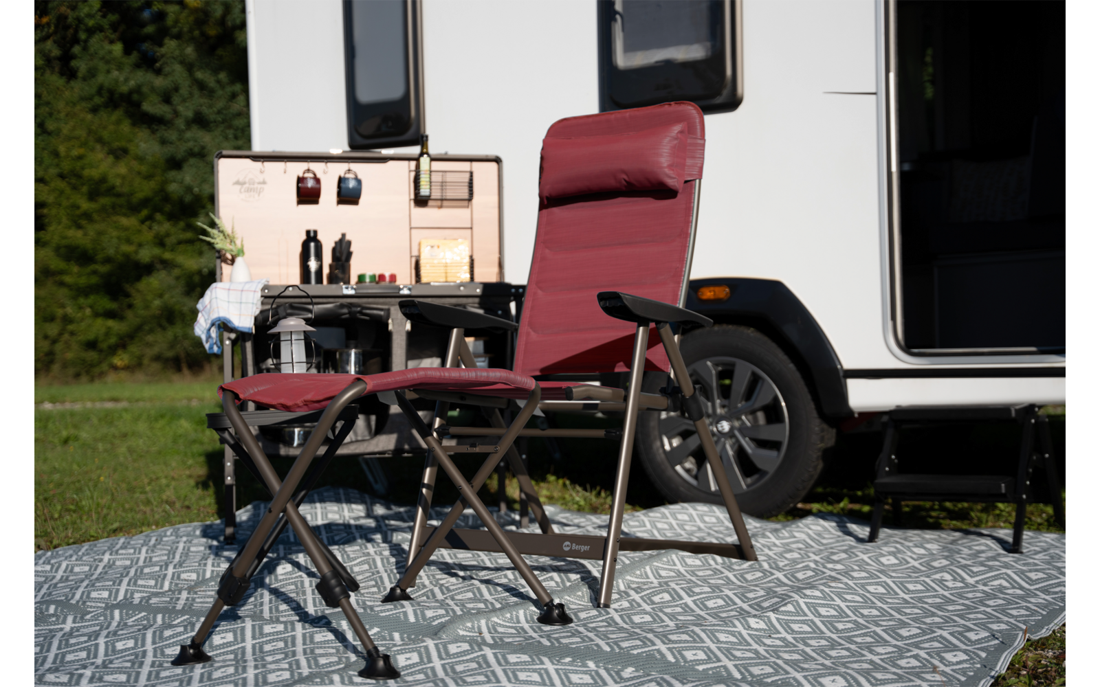 Zwei rote Klappstühle (ein hoher, ein niedriger Hocker) auf einem gemusterten Teppich vor einem weißen Wohnmobil; rechts ist ein Rad des Wohnmobils sichtbar, links ein kleiner Klapptisch mit offener Holzkiste mit Flaschen und Bechern, Gras und Bäume