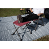 Person von Oberkörper bis Füße auf rotem klappbaren Campingstuhl, Beine auf passendem Fußhocker, weißes T‑Shirt, schwarze Hose, dunkle Sneakers; gemusteter Outdoor-Teppich, kleiner Beistelltisch mit Tasse, Gras und Autoreifen im Hintergrund