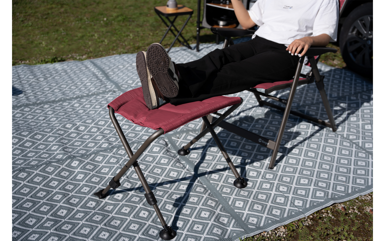 Person von Oberkörper bis Füße auf rotem klappbaren Campingstuhl, Beine auf passendem Fußhocker, weißes T‑Shirt, schwarze Hose, dunkle Sneakers; gemusteter Outdoor-Teppich, kleiner Beistelltisch mit Tasse, Gras und Autoreifen im Hintergrund