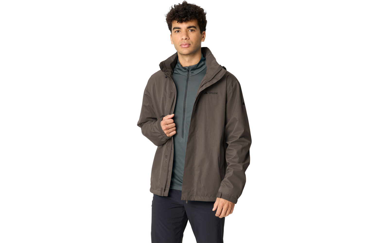 Mann mit dunklen, lockigen Haaren trägt eine graubraune Jacke über einem grünen Kapuzenpullover und schwarze Hose, steht vor weißem Hintergrund, hält die Jacke mit einer Hand