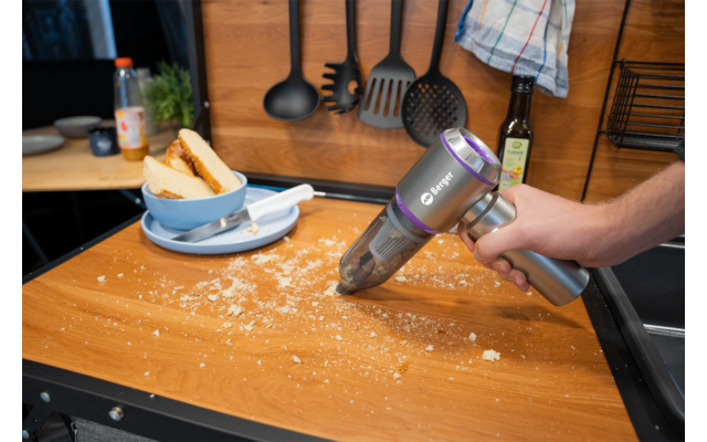 Main tenant un aspirateur à main sans fil au-dessus de miettes sur un plan de travail en bois ; assiette avec du pain, bols, bouteille d'huile, ustensiles de cuisine suspendus et torchon en arrière-plan