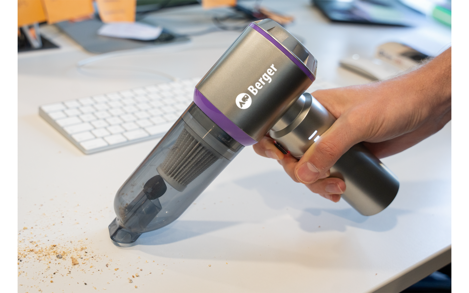 Main tenant un aspirateur à main sans fil portant la marque Berger, aspirant des miettes sur un bureau blanc à côté d'un clavier