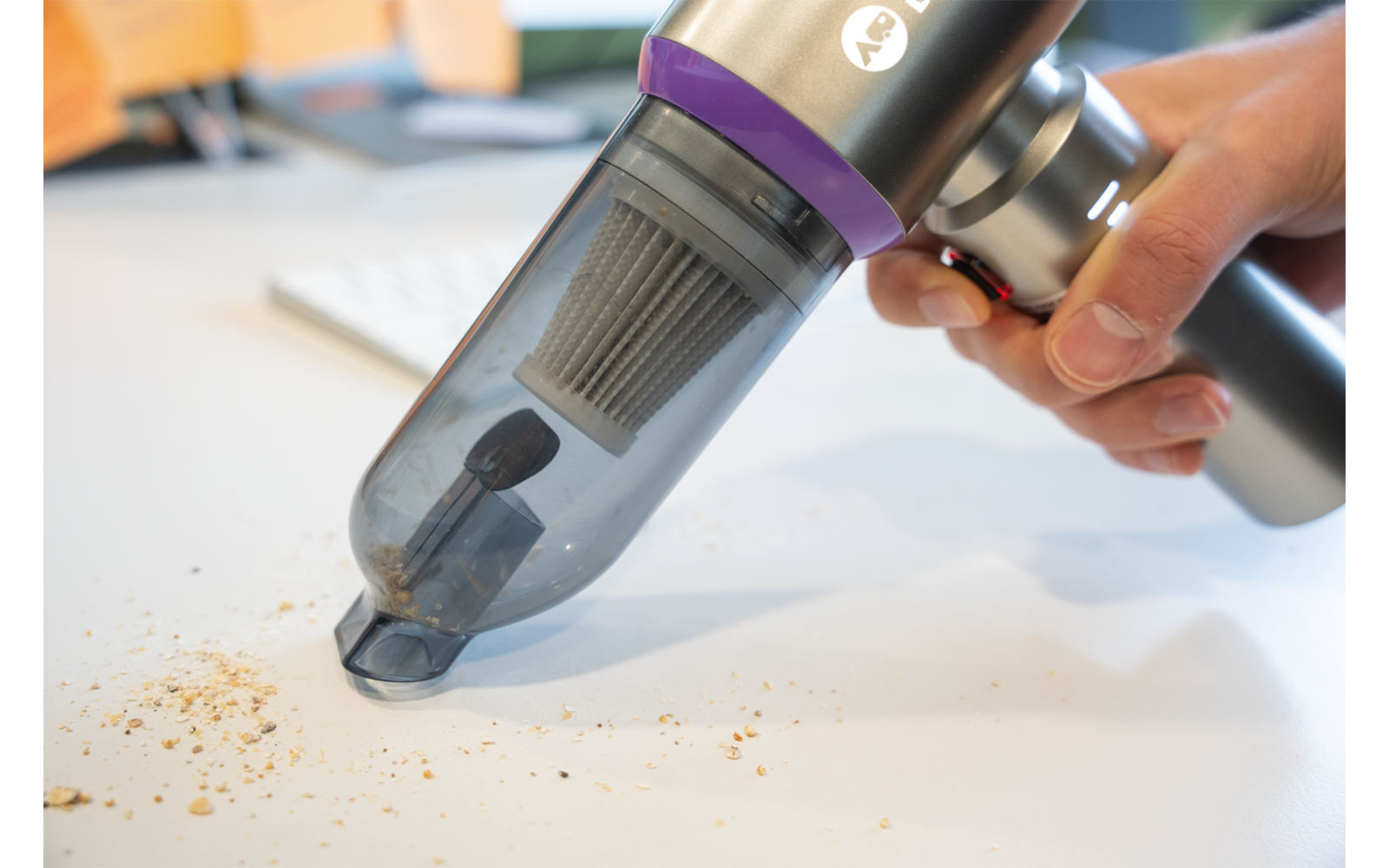 Main tenant un aspirateur à main sans fil au-dessus de miettes sur une surface blanche ; réservoir transparent avec filtre visible