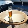 Halbvolles Pilsnerglas mit Schaum auf einer runden goldfarbenen Unterlage auf einem runden Holztisch; Bootsdeck und Wasser im Hintergrund