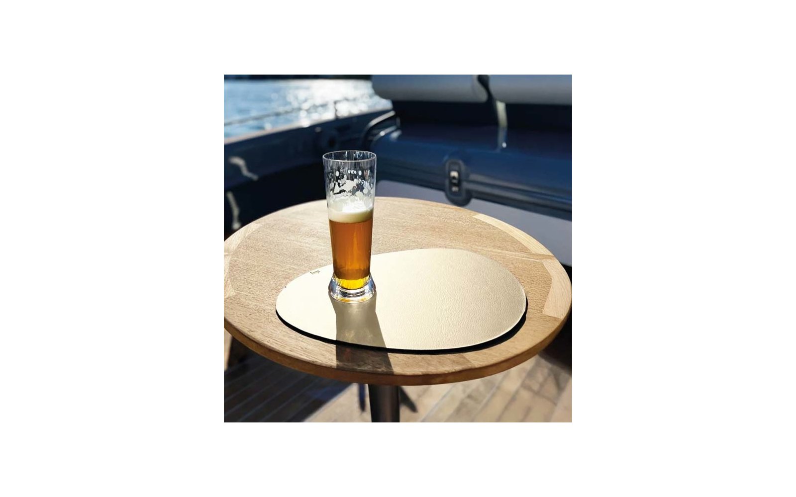 Halbvolles Pilsnerglas mit Schaum auf einer runden goldfarbenen Unterlage auf einem runden Holztisch; Bootsdeck und Wasser im Hintergrund