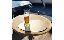 Verre de pils à moitié rempli avec mousse sur un dessous de verre rond couleur or posé sur une table ronde en bois ; pont de bateau et eau en arrière-plan