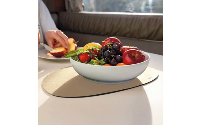 Weiße Schüssel mit Obst (rote Äpfel, dunkle Trauben, Erdbeeren, Orangen) auf ovaler hellbeiger Platzmatte auf einem hellen Tisch; im Hintergrund eine Hand, die einen Apfel auf einem kleinen Teller schneidet; gepolsterte Rückenlehne und Fenster mit Li