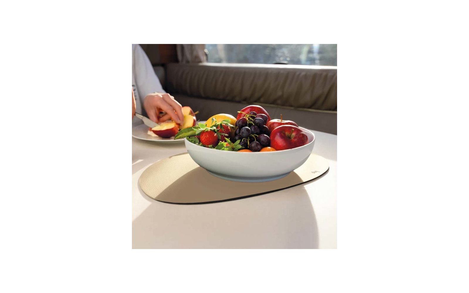 Weiße Schüssel mit Obst (rote Äpfel, dunkle Trauben, Erdbeeren, Orangen) auf ovaler hellbeiger Platzmatte auf einem hellen Tisch; im Hintergrund eine Hand, die einen Apfel auf einem kleinen Teller schneidet; gepolsterte Rückenlehne und Fenster mit Li