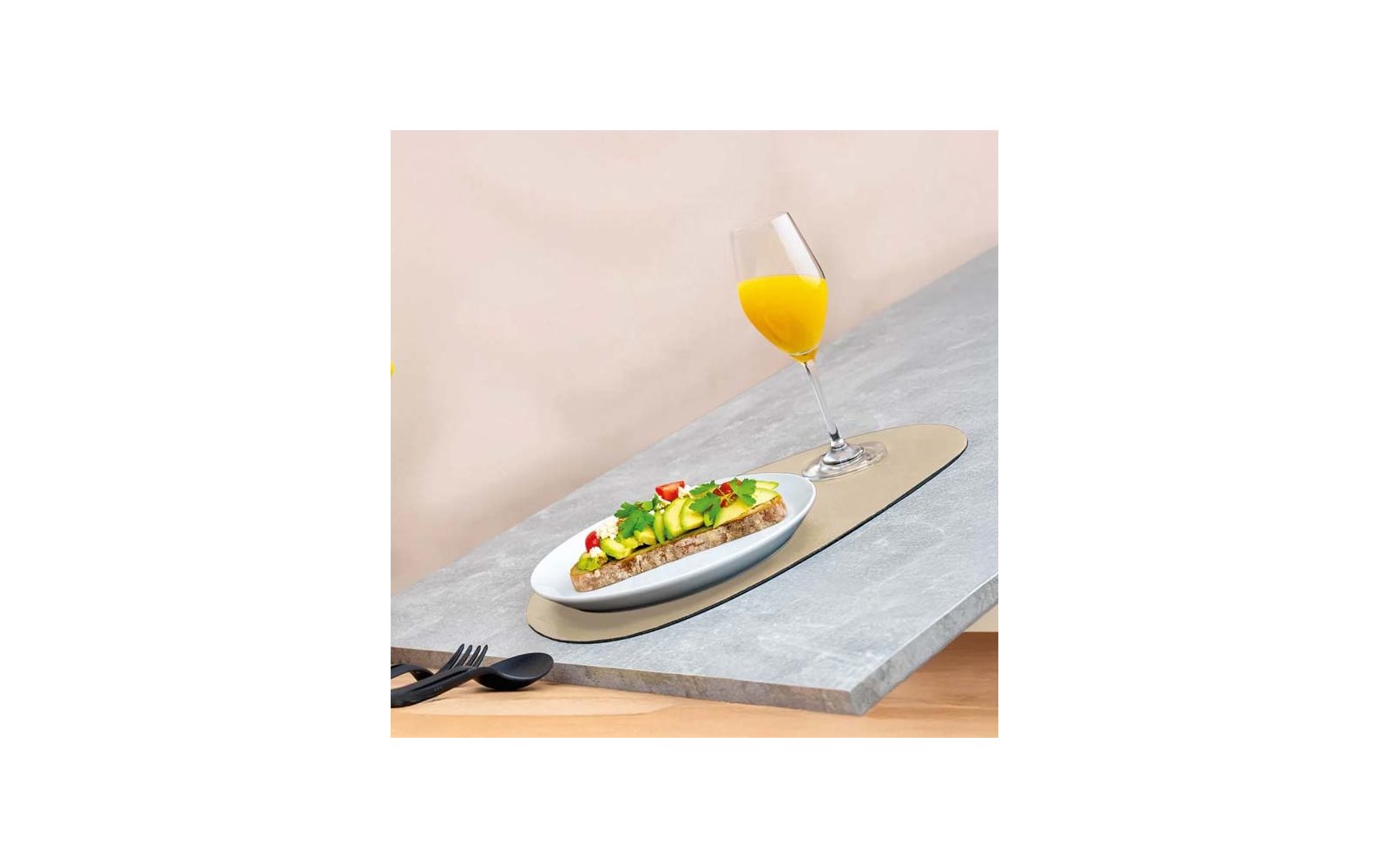 Scheibe Brot mit Samen, belegt mit Avocadoscheiben, halbierten Kirschtomaten und grünen Kräutern auf weißem Teller; Stielglas mit orangem Saft daneben; schwarze Gabel und Löffel auf grauem Tisch, beiger Hintergrund