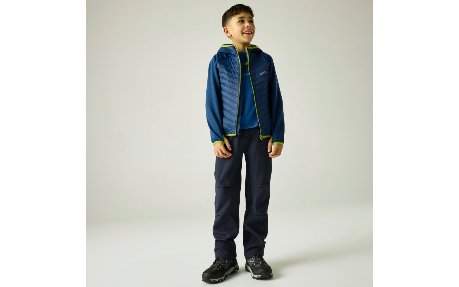 Junge steht, lächelnd, trägt dunkelblaue gesteppte Jacke mit hellgrünen Reißverschlusskanten, blaues T-Shirt, dunkle Hose und schwarze Sportschuhe, vor hellem neutralem Hintergrund