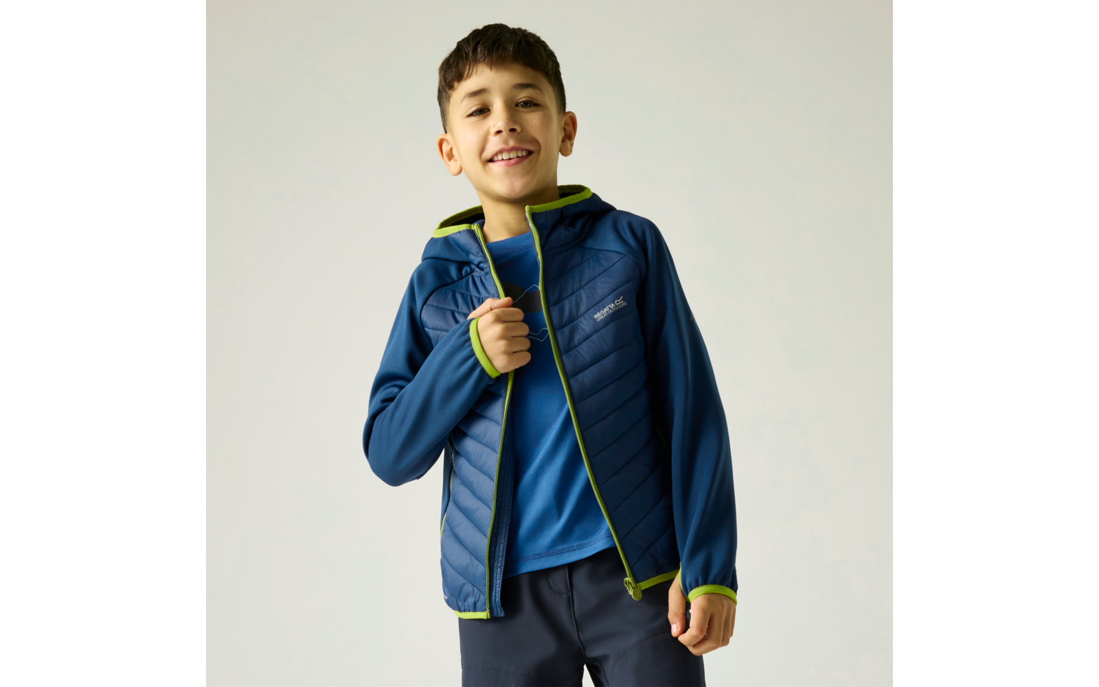 Ein lächelnder Junge in blauer gesteppter Jacke mit neongrünen Kanten über einem blauen T‑Shirt, hält den Reißverschlussrand, vor hellem Hintergrund