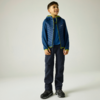 Junge steht, lächelnd, trägt dunkelblaue gesteppte Jacke mit hellgrünen Reißverschlusskanten, blaues T-Shirt, dunkle Hose und schwarze Sportschuhe, vor hellem neutralem Hintergrund