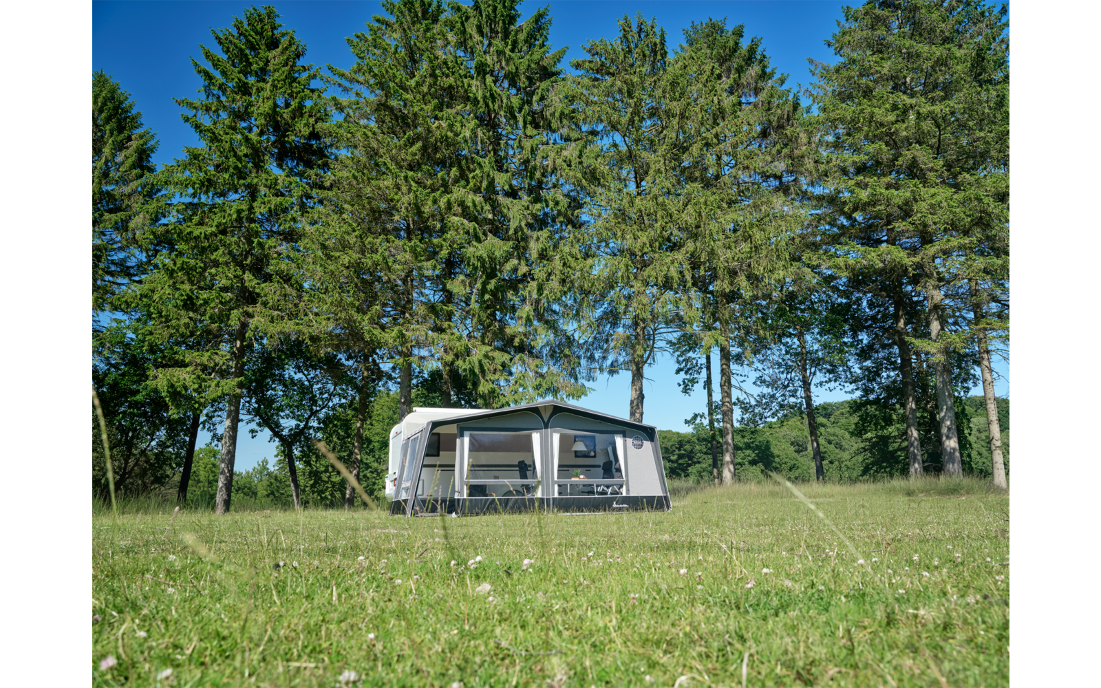 Weißer Wohnwagen mit angebautem Vorzelt auf grüner Wiese, im Hintergrund hohe Nadelbäume und blauer Himmel