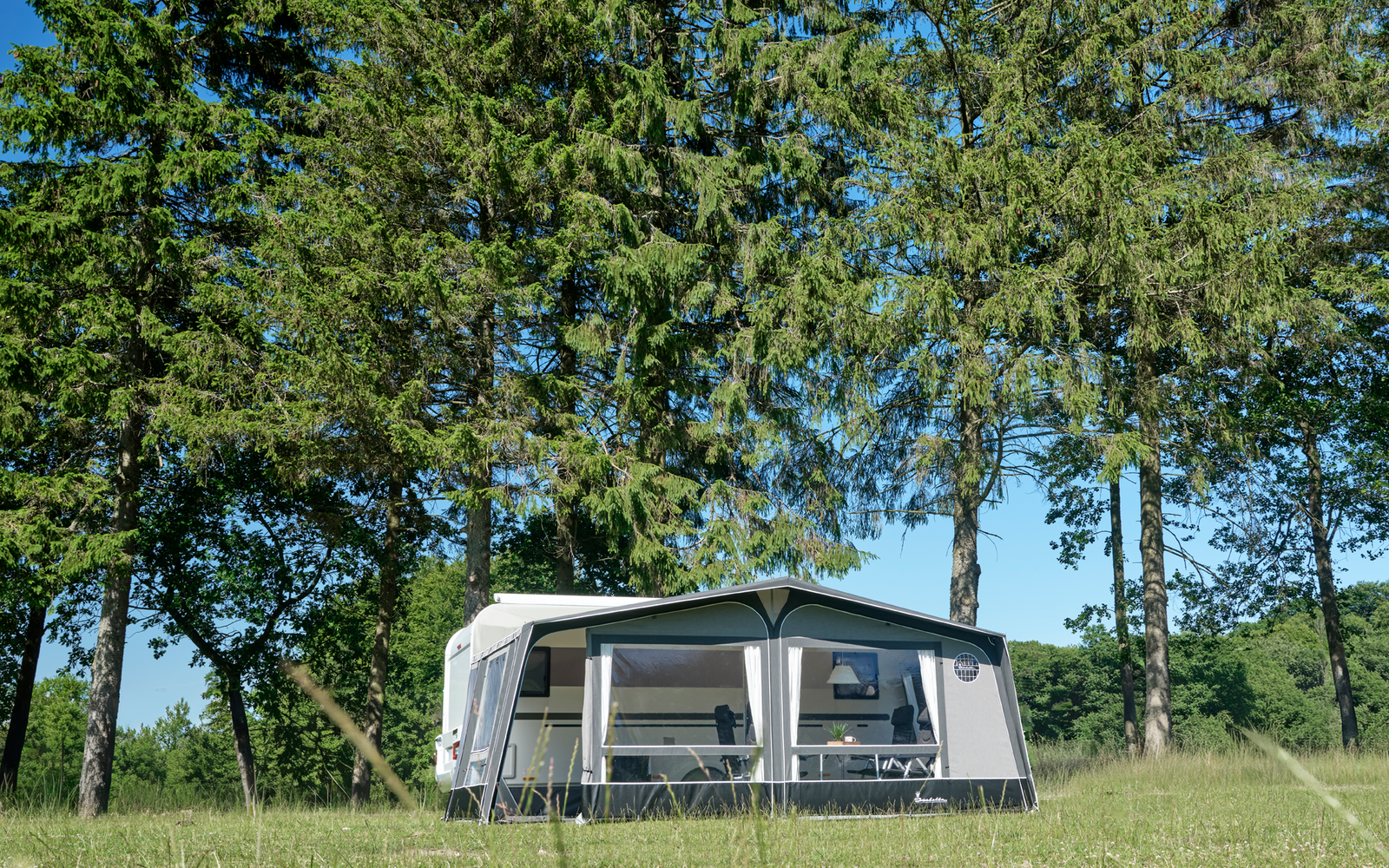 Weißer Wohnwagen mit angebautem Vorzelt auf grüner Wiese, im Hintergrund hohe Nadelbäume und blauer Himmel