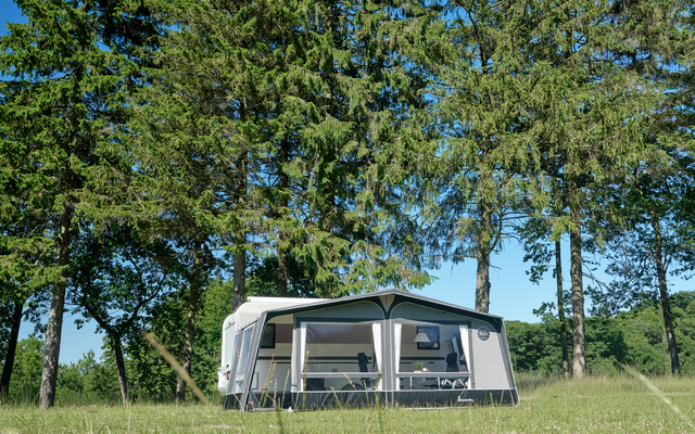 Weißer Wohnwagen mit angebautem Vorzelt auf grüner Wiese, im Hintergrund hohe Nadelbäume und blauer Himmel