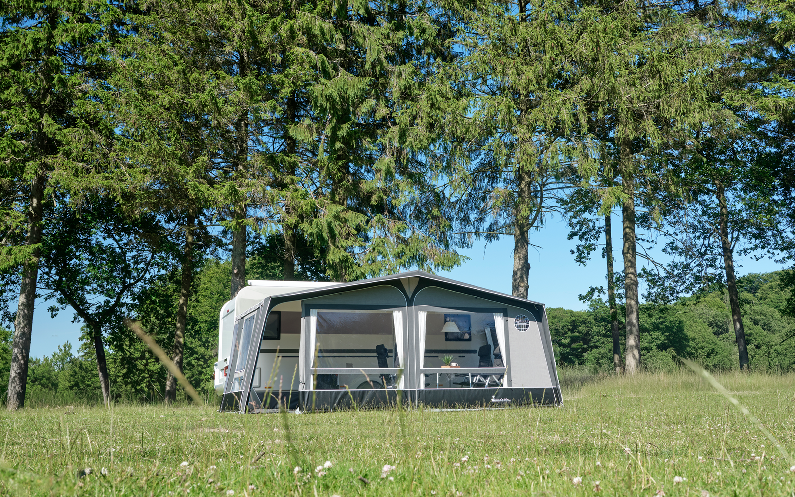 Weißer Wohnwagen mit angebautem Vorzelt auf grüner Wiese, im Hintergrund hohe Nadelbäume und blauer Himmel