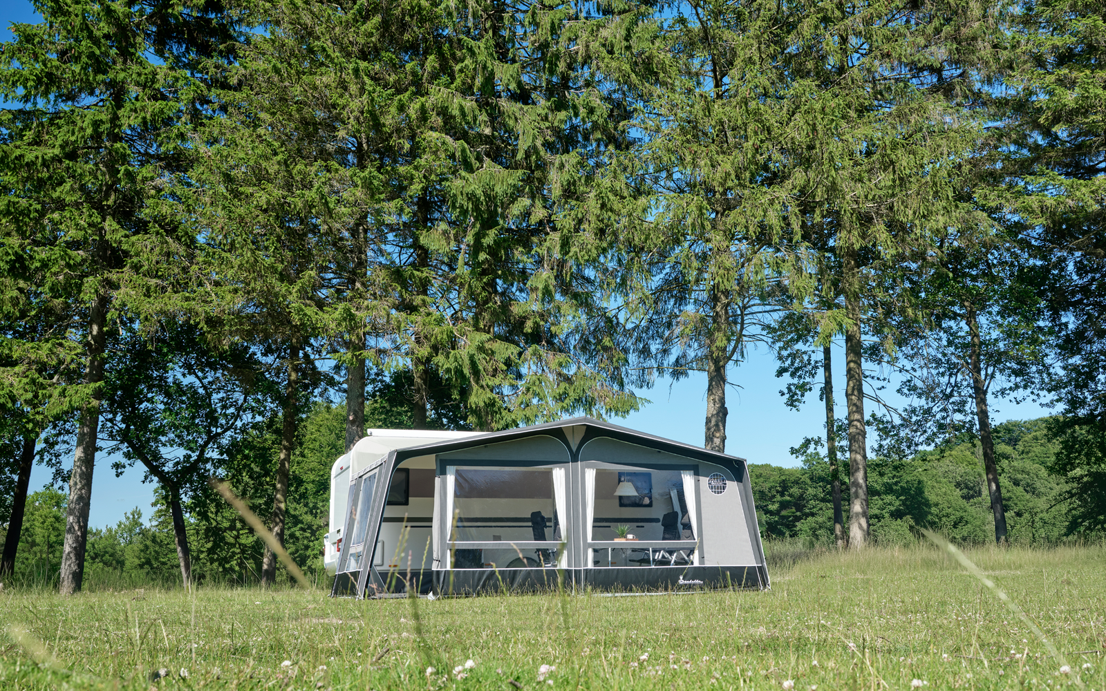 Caravana blanca con toldo anexo sobre un prado verde, al fondo altos árboles de hoja perenne y cielo azul