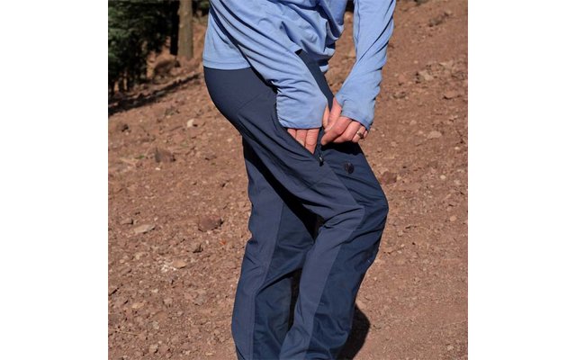 Unterkörper und Beine einer Person mit hellblauem Langarmshirt und dunkelblauen Hosen, eine Hand schließt den Reißverschluss einer seitlichen Hosentasche auf der Oberschenkelhöhe, steiniger brauner Boden im Hintergrund