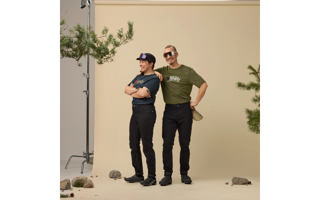 Twee personen voor een beige achtergrond; links een kleinere persoon met pet en gekruiste armen, rechts een langere persoon met zonnebril met één arm om de schouder van de ander; studiolichtstatief, dennetakken en stenen op de vloer zichtbaar