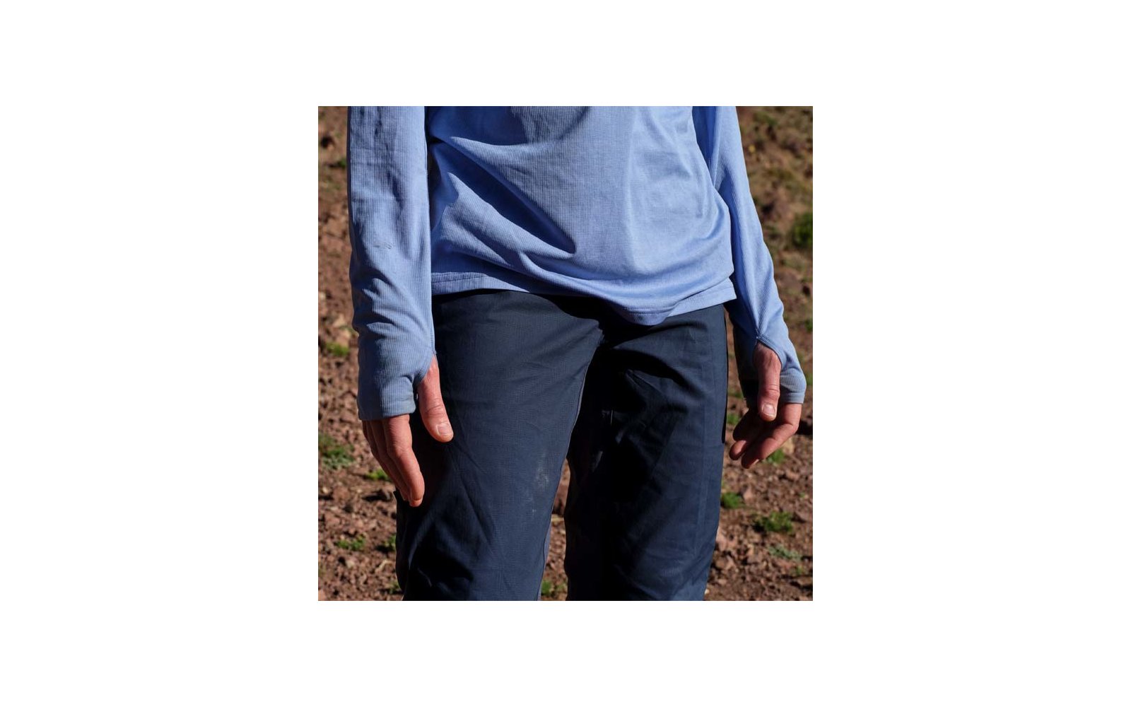 Oberkörper bis Oberschenkel einer Person in hellblauem Langarmshirt und dunkelblauen Hosen, Hände seitlich, trockener steiniger Boden im Hintergrund