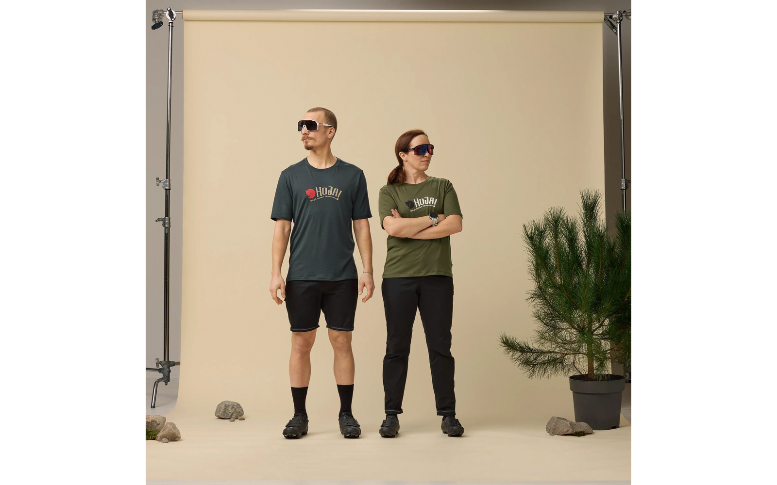 Twee personen met zonnebrillen staan naast elkaar voor een beige fotachtergrond. Links: donkergroen T‑shirt, zwarte short, zwarte sokken en zwarte schoenen. Rechts: olijfgroen T‑shirt, lange zwarte broek, zwarte schoenen, armen gekruist. Rechts een pot met een kleine den, meerdere stenen op de grond, achtergrondstatief zichtbaar