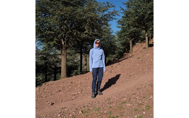 Person mit hellblauer Kapuzenjacke und Sonnenbrille steht auf einem schrägen, erdigen Hang vor hohen Nadelbäumen