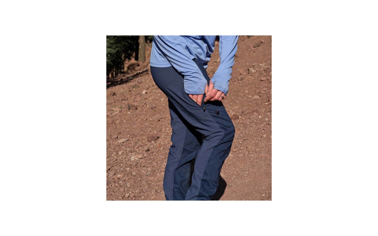 Unterkörper und Beine einer Person mit hellblauem Langarmshirt und dunkelblauen Hosen, eine Hand schließt den Reißverschluss einer seitlichen Hosentasche auf der Oberschenkelhöhe, steiniger brauner Boden im Hintergrund