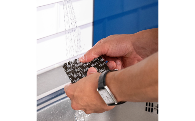 Deux mains tiennent une éponge noire avec un motif blanc sous l'eau courante au-dessus d'un évier ; à gauche des carreaux blancs et, en arrière-plan, un mur bleu