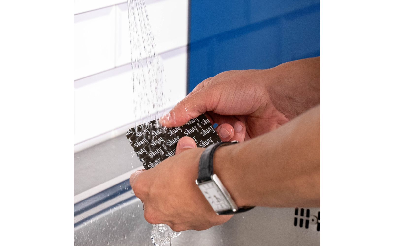 Deux mains tiennent une éponge noire avec un motif blanc sous l'eau courante au-dessus d'un évier ; à gauche des carreaux blancs et, en arrière-plan, un mur bleu