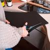 Mains en pull gris coupant avec des ciseaux une feuille noire sur un plan de travail de cuisine à côté d'une plaque vitrocéramique ; règle, verres et petites plantes visibles