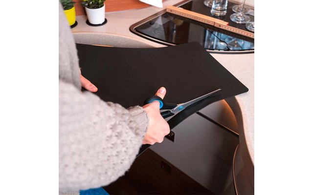 Mains en pull gris coupant avec des ciseaux une feuille noire sur un plan de travail de cuisine à côté d'une plaque vitrocéramique ; règle, verres et petites plantes visibles