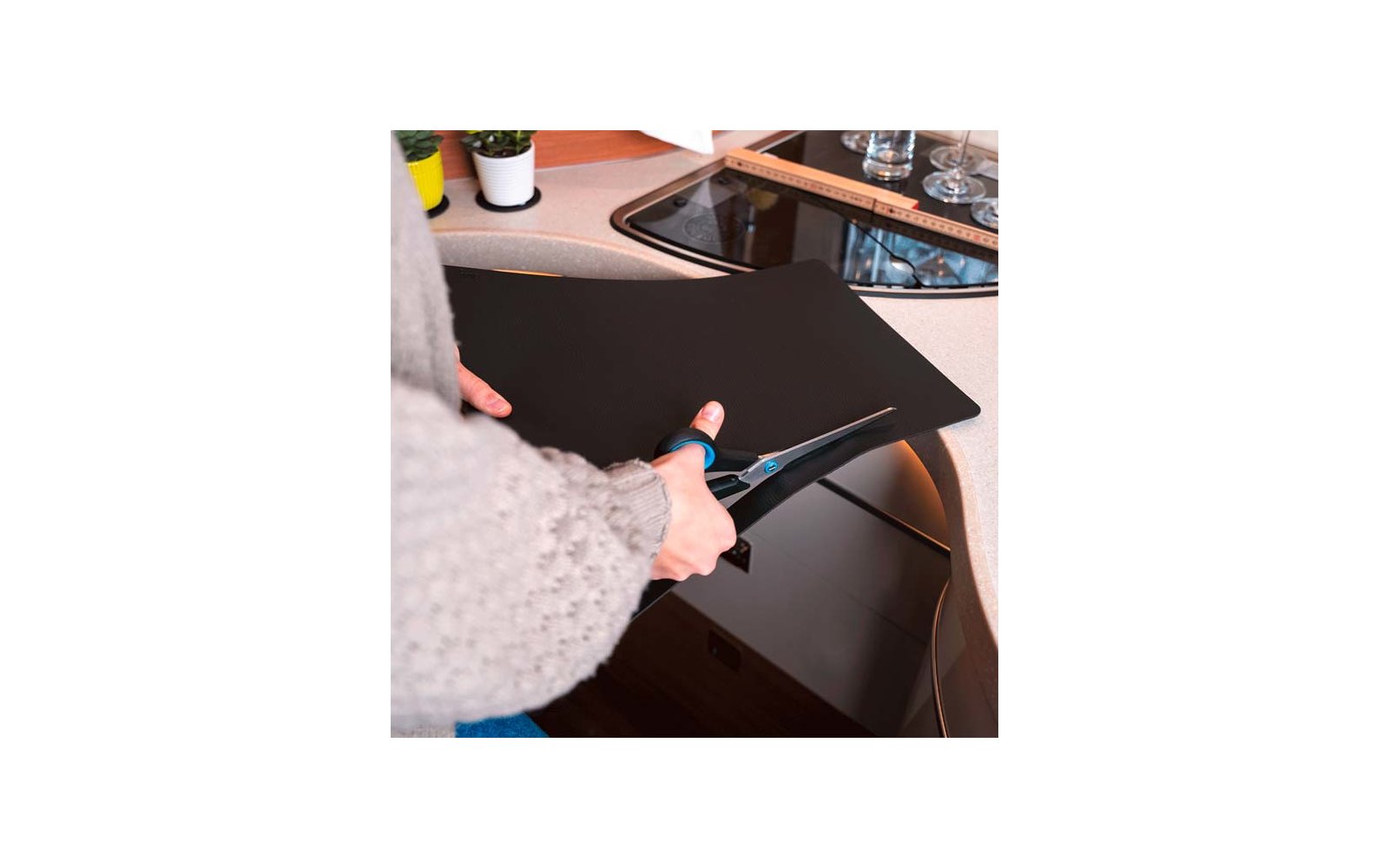 Mains en pull gris coupant avec des ciseaux une feuille noire sur un plan de travail de cuisine à côté d'une plaque vitrocéramique ; règle, verres et petites plantes visibles