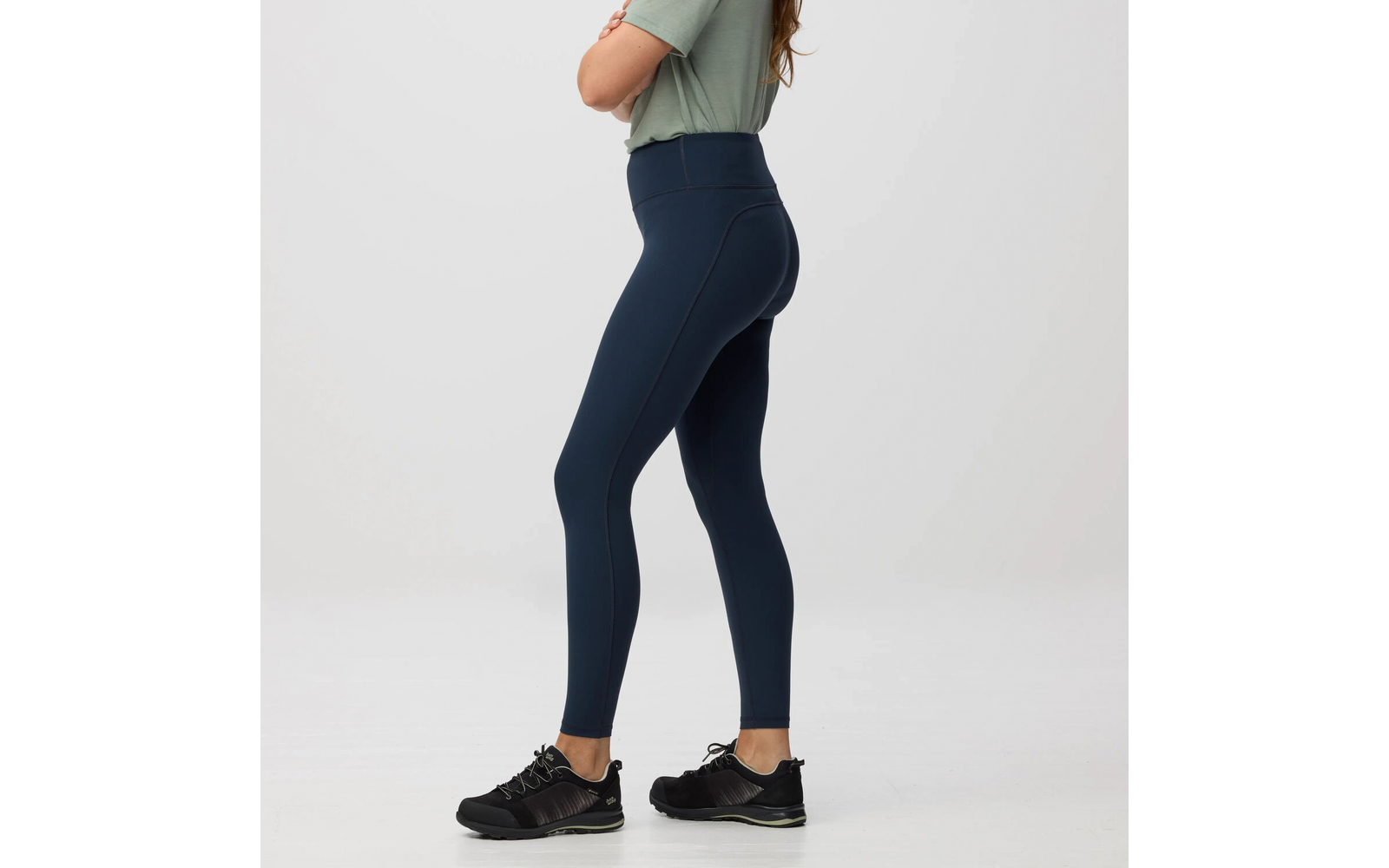 Persona de perfil desde los hombros hasta los pies, con camiseta verde claro, mallas azul oscuro de talle alto y zapatillas deportivas negras sobre fondo claro