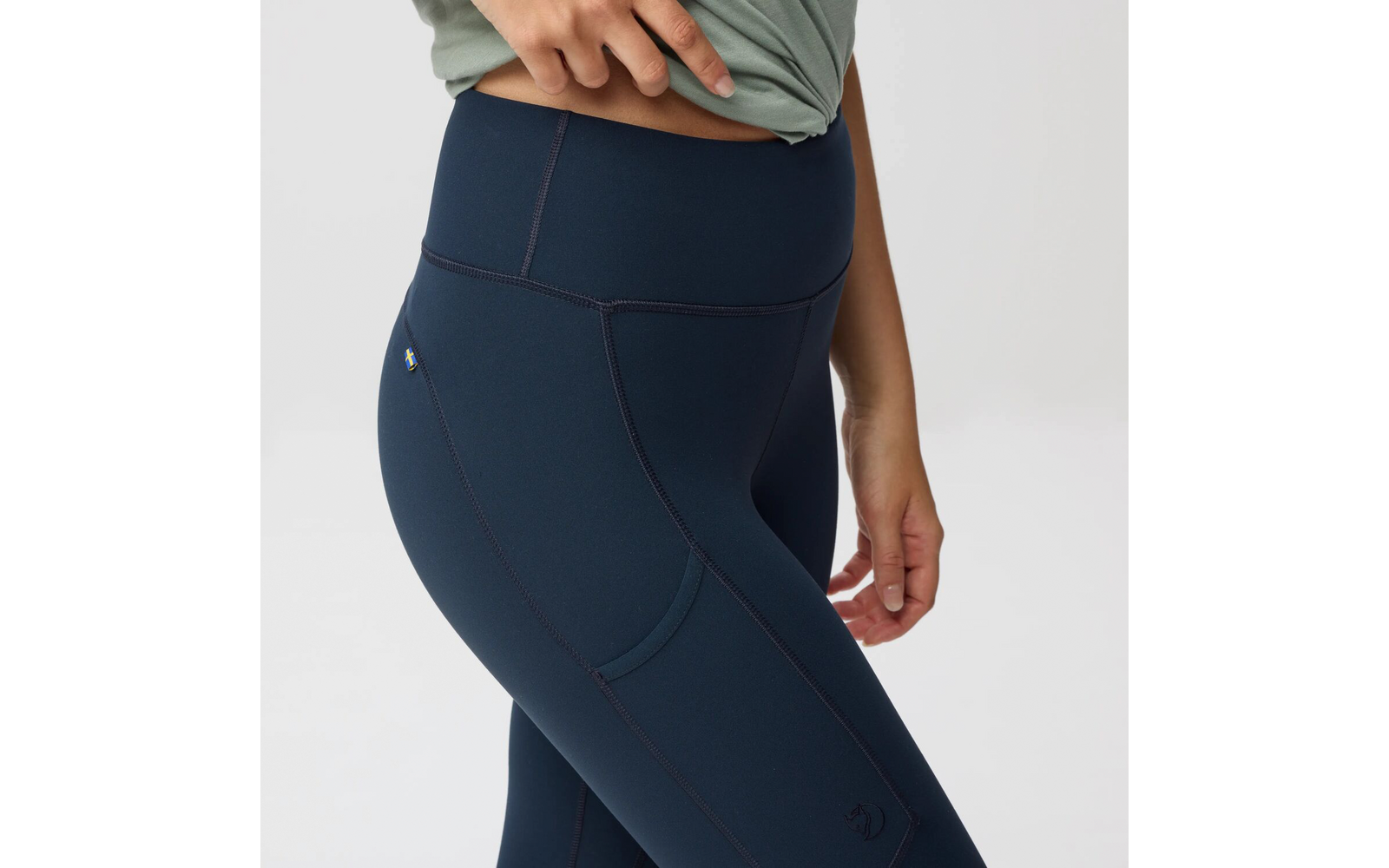 Cadera y parte superior de los muslos de una persona con mallas azul oscuro de talle alto, bolsillo lateral y pequeño logo; mano derecha levanta ligeramente una prenda superior, abdomen inferior visible