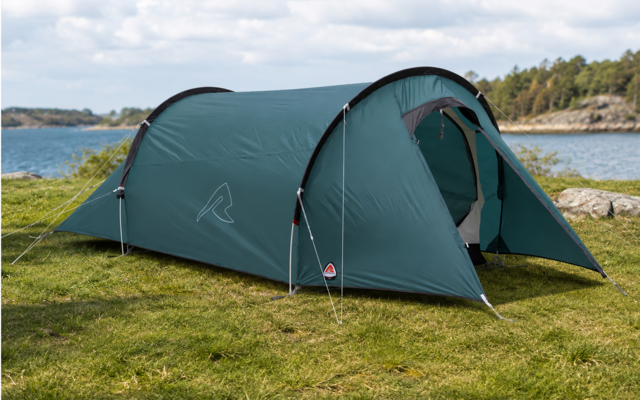 Green tunnel tent with open entrance on grass at a lakeshore; tent poles visible, rocks and trees on the opposite shore