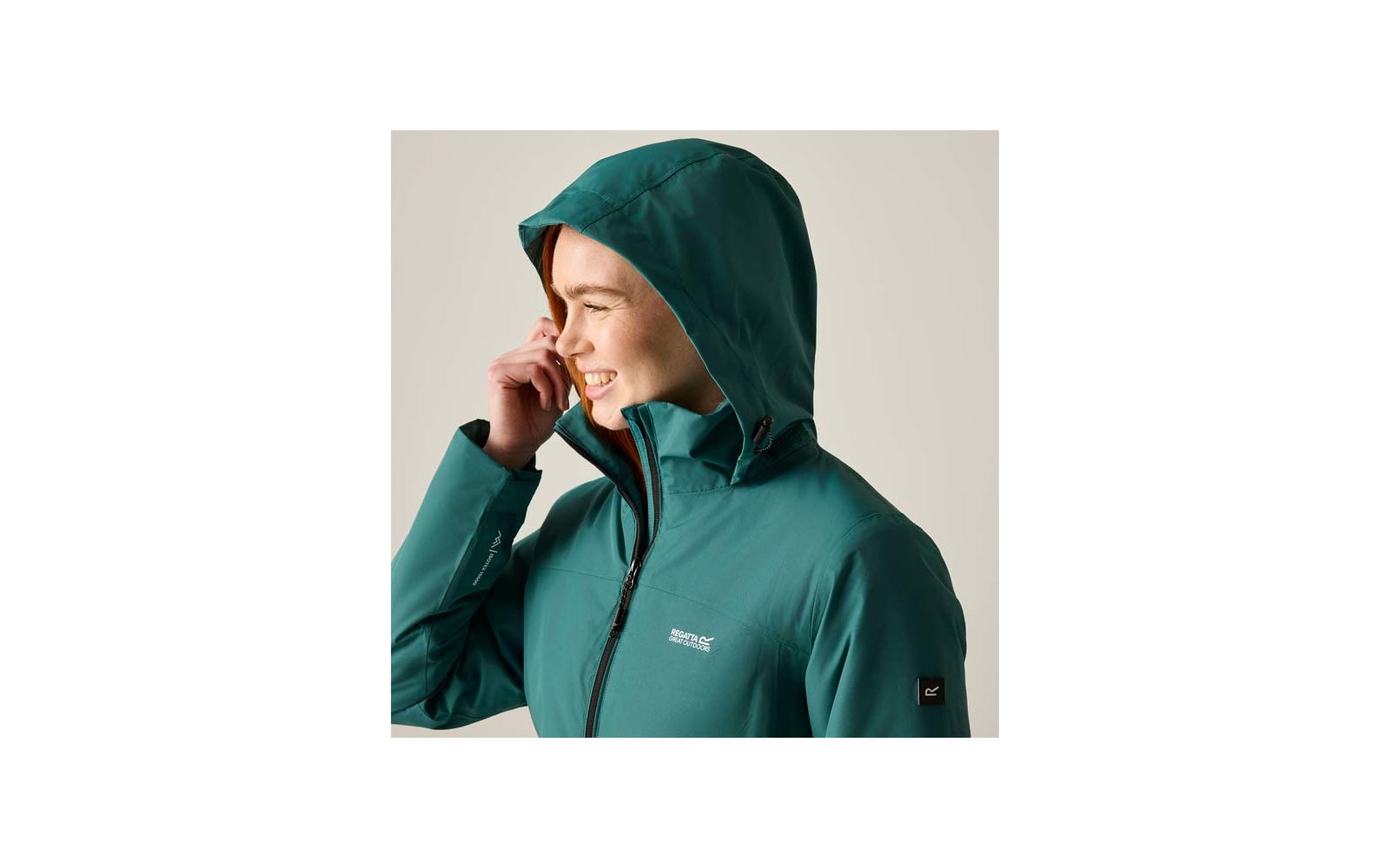 Person in profile, smiling with hand near face, wearing green hooded jacket with zipper and logo, light background