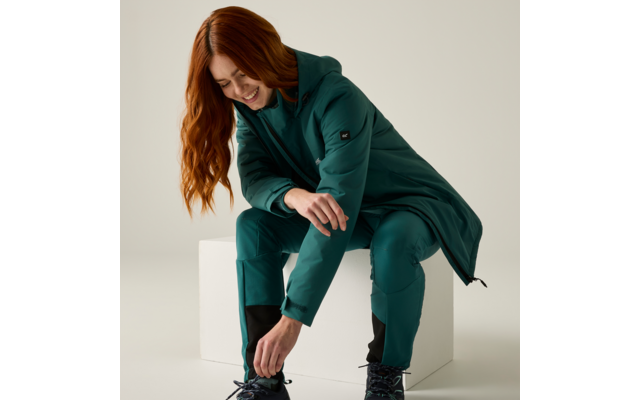 Person with long red hair sitting on a white cube, wearing a green coat and trousers, tying a shoelace