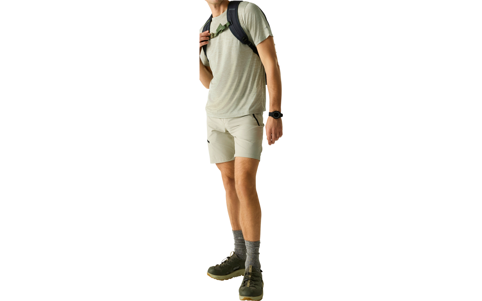 Mann steht vor hellem Hintergrund, trägt graues T‑Shirt, helle Shorts, graue Socken und grüne Schuhe, Rucksack über einer Schulter, Armbanduhr, Blick nach rechts