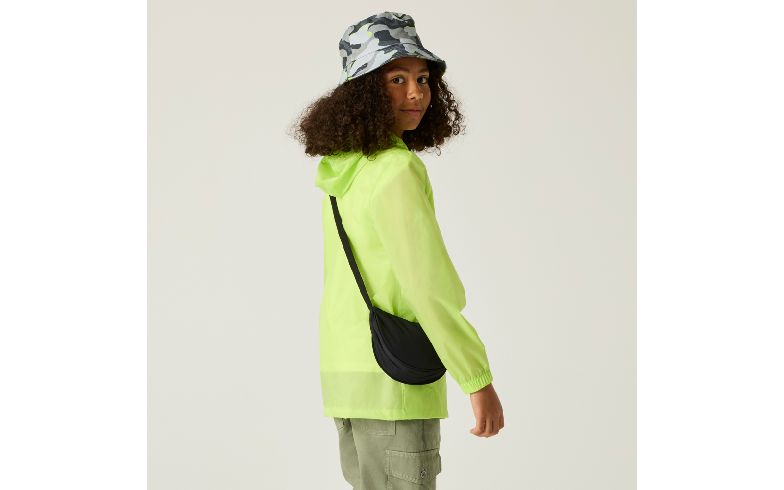 Person mit lockigem Haar, trägt camo-Bucket-Hut, neon-gelbe transparente Kapuzenjacke, olivgrüne Cargohose und schwarze Umhängetasche, schaut über die Schulter vor hellem Hintergrund