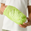 Hände halten eine hellgrüne, kompakt verpackte Kinderjacke in einem Beutel mit weißer Aufschrift und Logo; weißes T-Shirt im Hintergrund