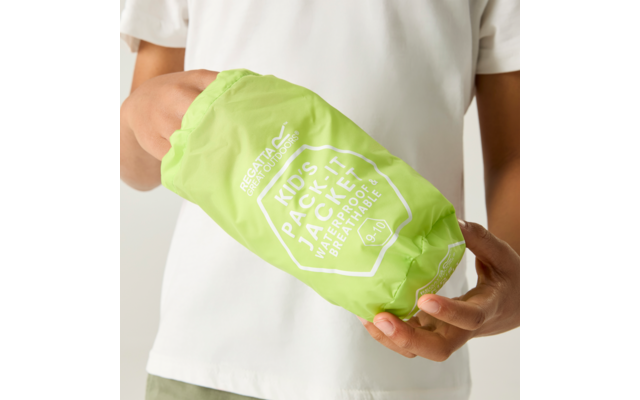 Hände halten eine hellgrüne, kompakt verpackte Kinderjacke in einem Beutel mit weißer Aufschrift und Logo; weißes T-Shirt im Hintergrund