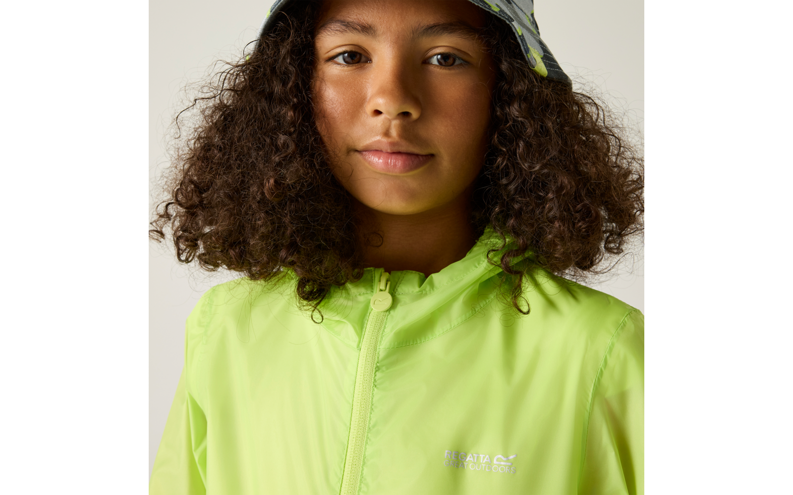 Nahaufnahme einer Person mit lockigen Haaren, einer neongrünen Regenjacke mit Reißverschluss und einem gemusterten Fischerhut vor hellem Hintergrund