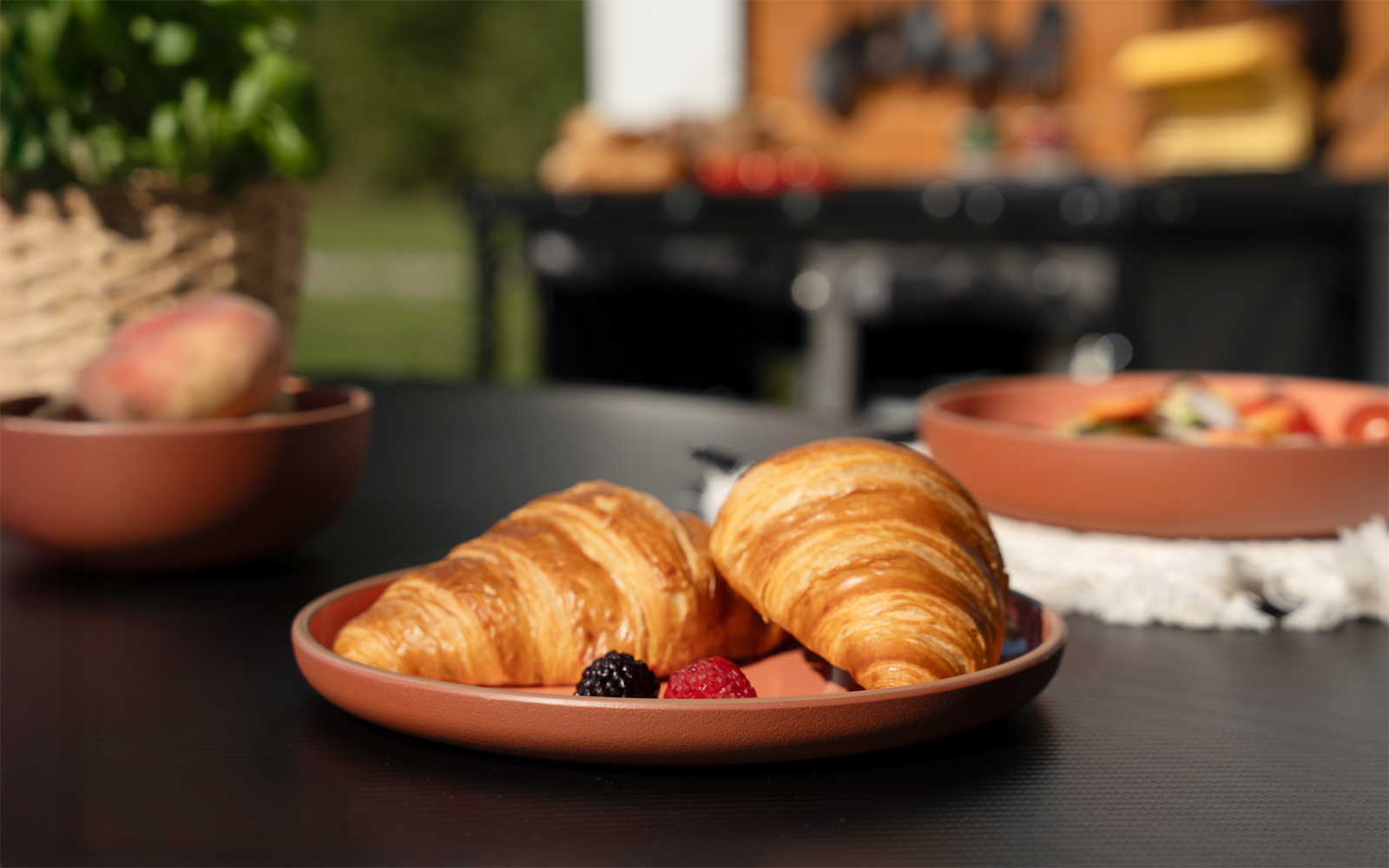 Zwei Croissants auf einem runden Terrakotta-Teller auf einem schwarzen Tisch; drei Beeren vorn; im Hintergrund unscharfe Schalen und ein Korb